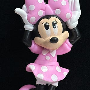 May include: A necklace pendant featuring a cartoon mouse character wearing a pink dress with white polka dots and a matching bow. The character has white gloves and pink shoes. The necklace chain consists of alternating pink and clear beads.