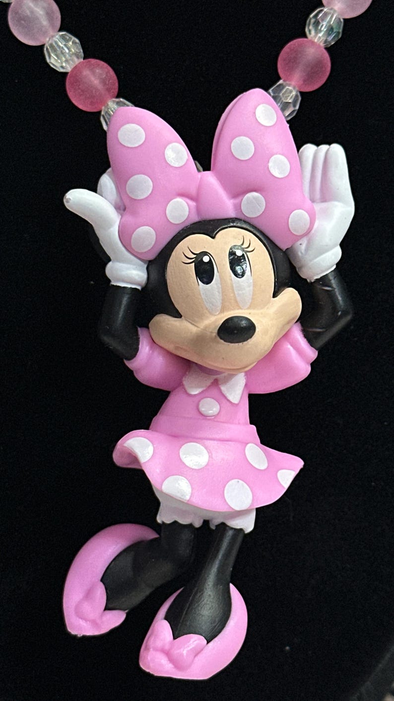 May include: A necklace pendant featuring a cartoon mouse character wearing a pink dress with white polka dots and a matching bow. The character has white gloves and pink shoes. The necklace chain consists of alternating pink and clear beads.
