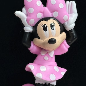 May include: A necklace pendant featuring a cartoon mouse character wearing a pink dress with white polka dots and a matching bow. The character has white gloves and pink shoes. The necklace strand consists of alternating pink and clear beads.