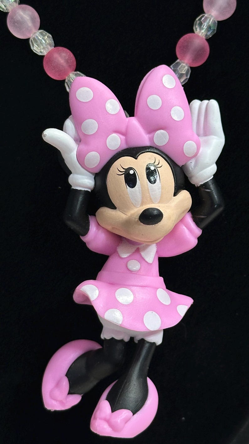 May include: A necklace pendant featuring a cartoon mouse character wearing a pink dress with white polka dots and a matching bow. The character has white gloves and pink shoes. The necklace strand consists of alternating pink and clear beads.