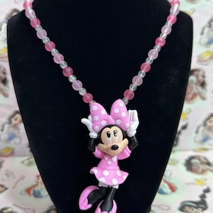 May include: A necklace featuring a Minnie Mouse pendant with a pink polka dot dress and bow, white gloves, and black shoes. The chain is composed of alternating translucent clear and opaque pink beads, creating a colorful and playful accessory.