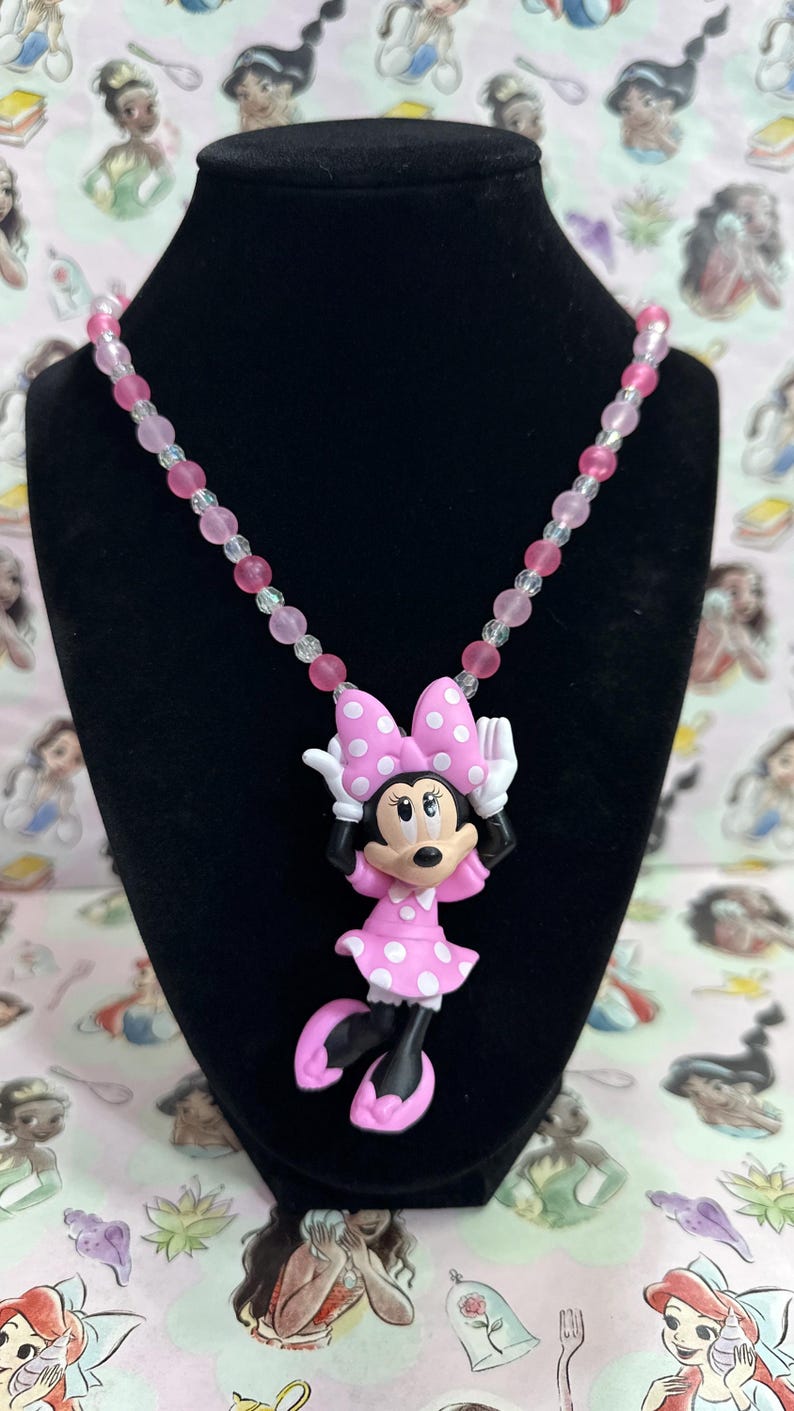 May include: A necklace featuring a Minnie Mouse pendant with a pink polka dot dress and bow, white gloves, and black shoes. The chain is composed of alternating translucent clear and opaque pink beads, creating a colorful and playful accessory.