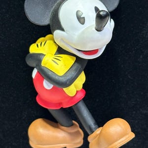 Puede incluir: Un collar con un colgante de Mickey Mouse. El colgante presenta a Mickey Mouse con su atuendo cl&aacute;sico: pantalones cortos rojos, guantes amarillos y zapatos marrones. El collar tiene una cadena de cuentas negras, amarillas y rojas. El fondo es negro.