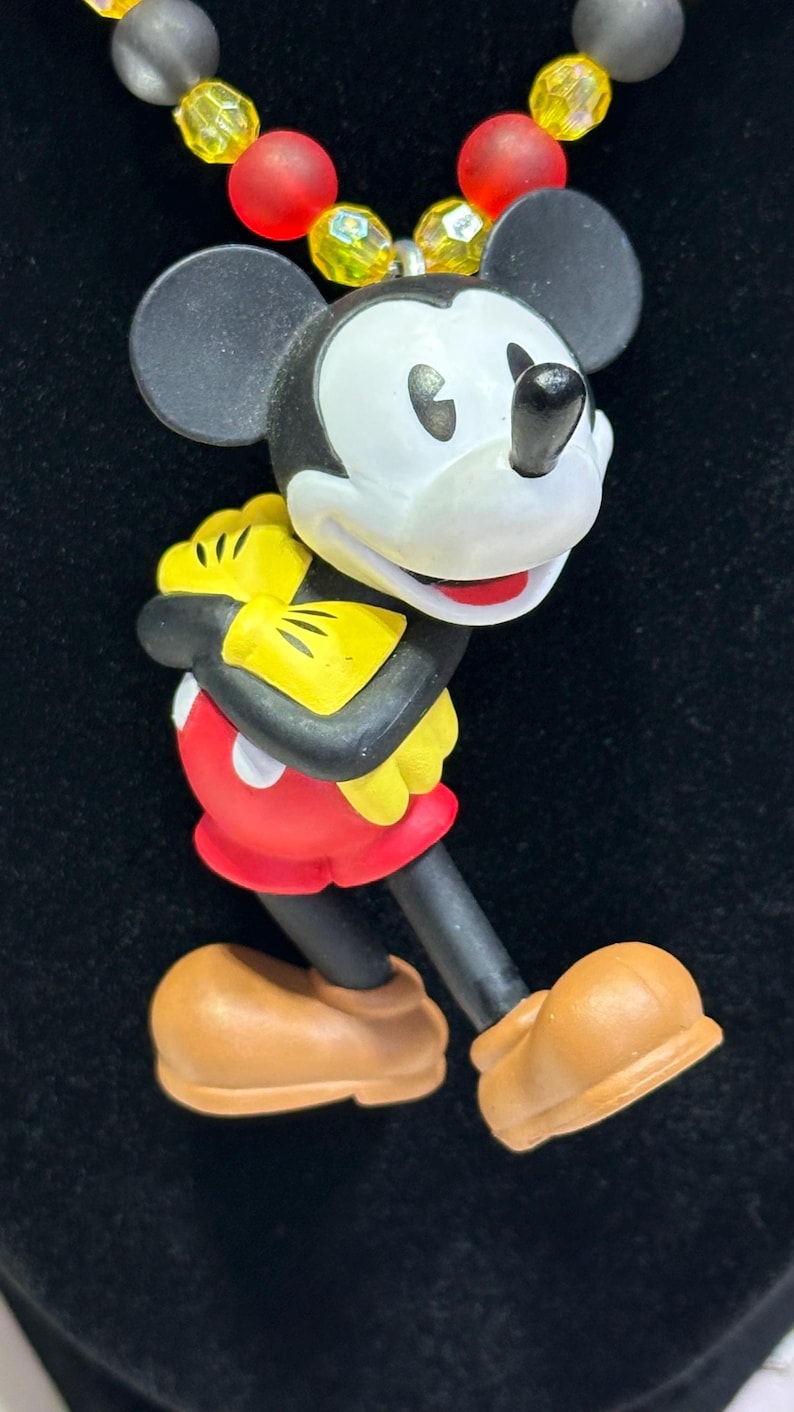 Puede incluir: Un collar con un colgante de Mickey Mouse. El colgante presenta a Mickey Mouse con su atuendo cl&aacute;sico: pantalones cortos rojos, guantes amarillos y zapatos marrones. El collar tiene una cadena de cuentas negras, amarillas y rojas. El fondo es negro.