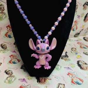 K&ouml;nnte beinhalten: Eine Halskette mit einer Kette aus hellvioletten und rosafarbenen Perlen und einem zentralen Anh&auml;nger in Form einer Zeichentrickfigur mit gro&szlig;en Ohren, kleiner Nase und hellviolettem K&ouml;rper, die einen hellrosa Brustfleck aufweist.