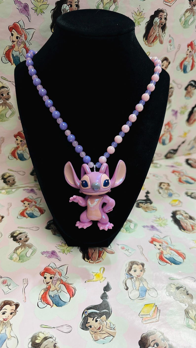 K&ouml;nnte beinhalten: Eine Halskette mit einer Kette aus hellvioletten und rosafarbenen Perlen und einem zentralen Anh&auml;nger in Form einer Zeichentrickfigur mit gro&szlig;en Ohren, kleiner Nase und hellviolettem K&ouml;rper, die einen hellrosa Brustfleck aufweist.