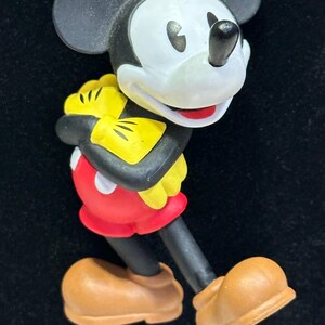 Puede incluir: Un colgante de collar con la figura de Mickey Mouse. El colgante presenta a Mickey Mouse con su atuendo cl&aacute;sico: pantalones cortos rojos, guantes amarillos y zapatos marrones. El collar tiene un cord&oacute;n de cuentas con cuentas rojas, amarillas y grises. El fondo es negro.