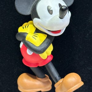 Puede incluir: Un colgante de Mickey Mouse con cuerpo negro, cara blanca y pantalones cortos rojos. El colgante forma parte de un collar con un hilo de cuentas amarillas, rojas y grises. El personaje de dibujos animados sostiene un objeto amarillo.