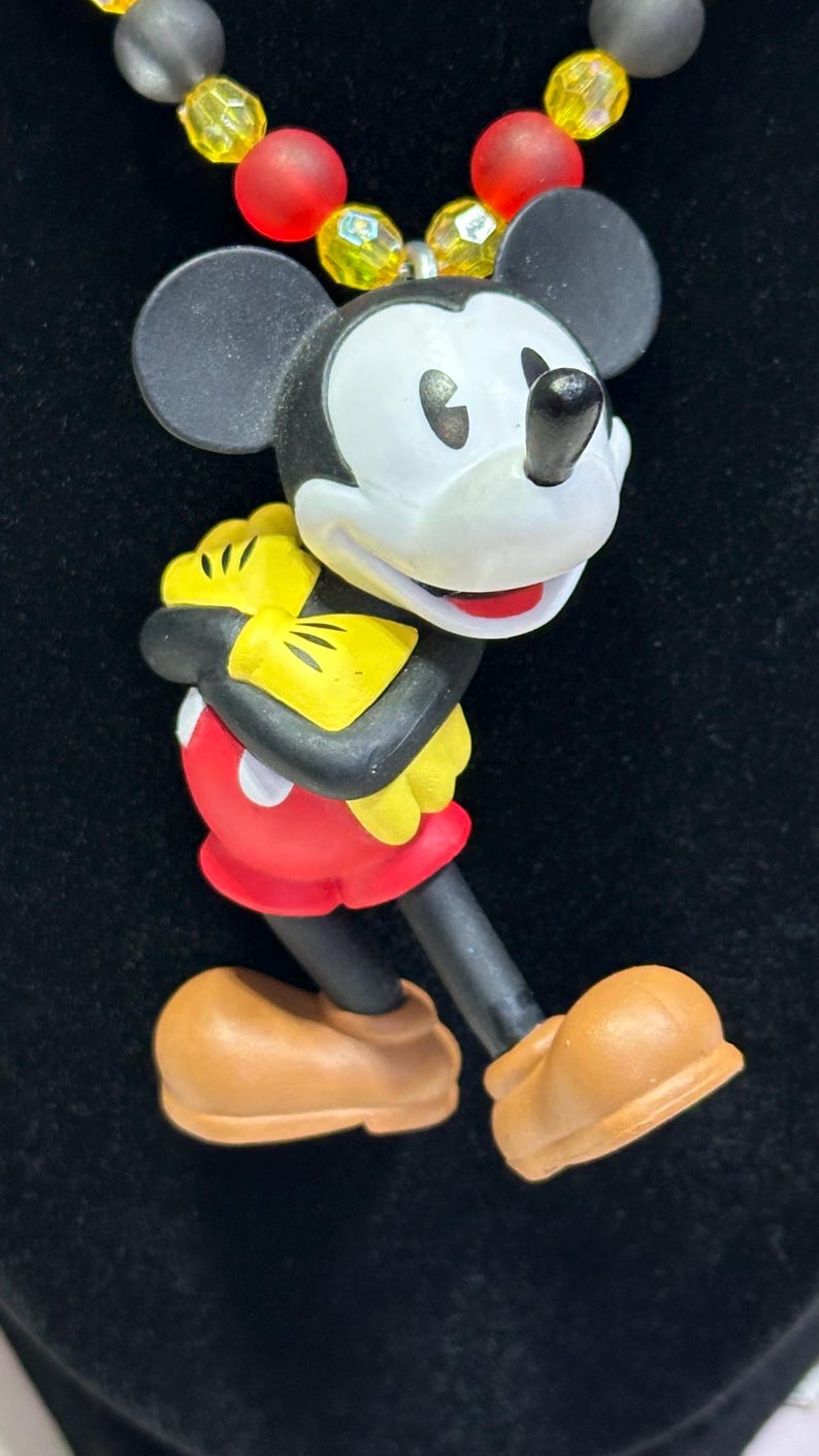 Puede incluir: Un colgante de Mickey Mouse con cuerpo negro, cara blanca y pantalones cortos rojos. El colgante forma parte de un collar con un hilo de cuentas amarillas, rojas y grises. El personaje de dibujos animados sostiene un objeto amarillo.