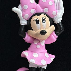 May include: A necklace featuring a Minnie Mouse pendant with a pink polka dot bow and dress, white gloves, and black shoes. The pendant is attached to a beaded chain made of alternating translucent clear and matte pink spherical beads.