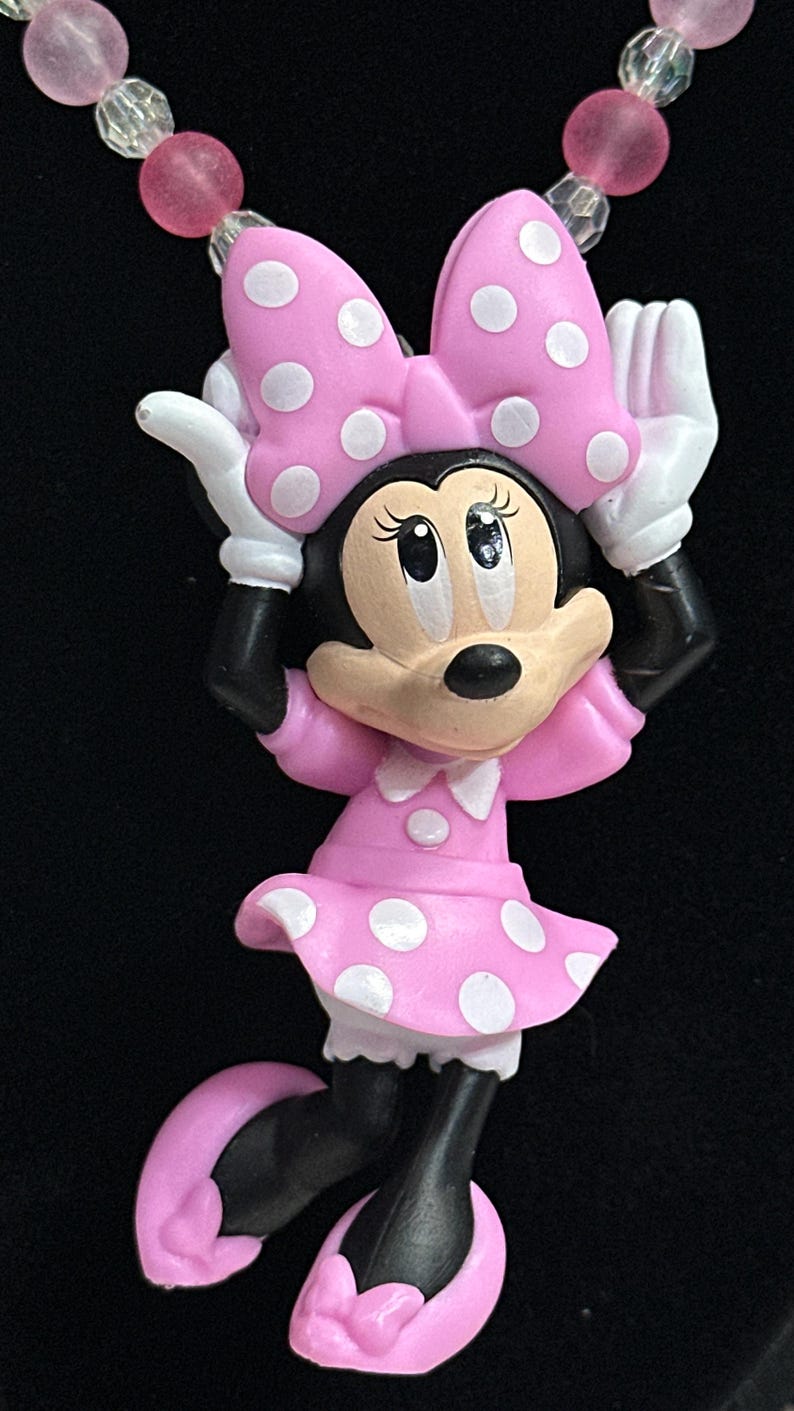 May include: A necklace featuring a Minnie Mouse pendant with a pink polka dot bow and dress, white gloves, and black shoes. The pendant is attached to a beaded chain made of alternating translucent clear and matte pink spherical beads.