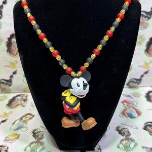 Puede incluir: Un collar con colgante de Mickey Mouse. El collar presenta un dije de Mickey Mouse con cuentas rojas, amarillas y negras. El collar se exhibe en un maniqu&iacute; negro. El fondo es un patr&oacute;n rosa con ilustraciones de princesas de Disney.