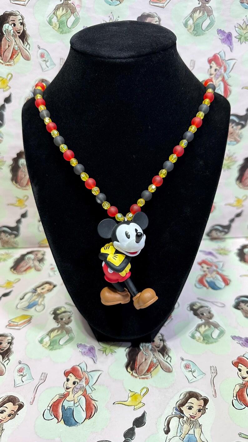 Puede incluir: Un collar con colgante de Mickey Mouse. El collar presenta un dije de Mickey Mouse con cuentas rojas, amarillas y negras. El collar se exhibe en un maniqu&iacute; negro. El fondo es un patr&oacute;n rosa con ilustraciones de princesas de Disney.
