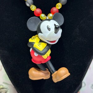 Puede incluir: Un collar con colgante de Mickey Mouse. El colgante presenta a Mickey con su atuendo cl&aacute;sico, con pantalones cortos rojos, guantes amarillos y zapatos marrones. El collar tiene una cadena de cuentas con cuentas rojas, amarillas y negras. El collar se exhibe sobre una superficie negra.