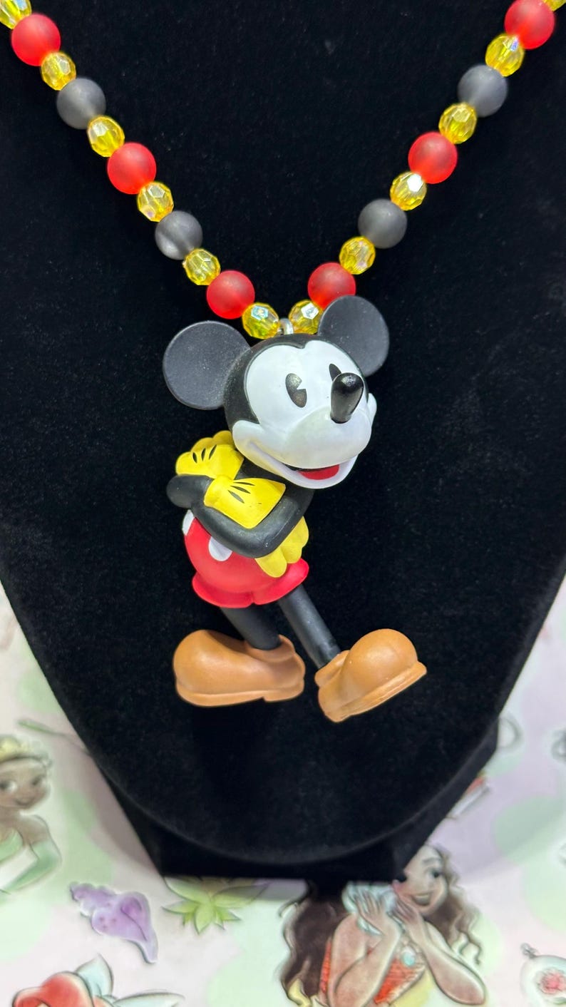 Puede incluir: Un collar con colgante de Mickey Mouse. El colgante presenta a Mickey con su atuendo cl&aacute;sico, con pantalones cortos rojos, guantes amarillos y zapatos marrones. El collar tiene una cadena de cuentas con cuentas rojas, amarillas y negras. El collar se exhibe sobre una superficie negra.