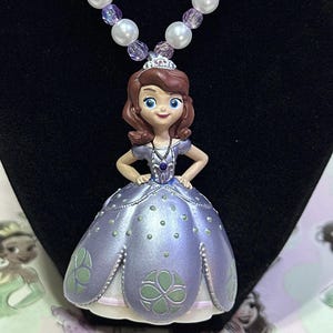 May include: A necklace featuring a pendant of a princess in a lavender dress, with a tiara and a beaded necklace. The necklace has purple and white beads. The princess pendant has brown hair and blue eyes. The necklace is displayed on a black background.