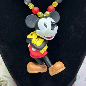 Puede incluir: Un collar con colgante de Mickey Mouse. El colgante es una figura de dibujos animados de Mickey Mouse con pantalones cortos rojos, guantes amarillos y zapatos marrones. El collar tiene una hebra de cuentas rojas, amarillas y grises.