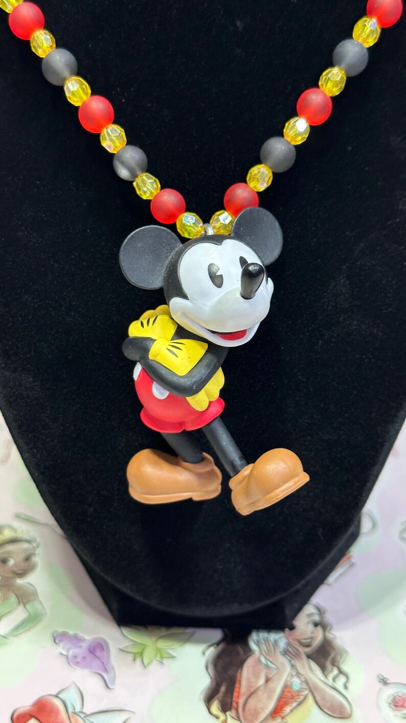 Puede incluir: Un collar con colgante de Mickey Mouse. El colgante es una figura de dibujos animados de Mickey Mouse con pantalones cortos rojos, guantes amarillos y zapatos marrones. El collar tiene una hebra de cuentas rojas, amarillas y grises.