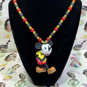 Puede incluir: Un collar con un colgante de Mickey Mouse. La cadena de cuentas tiene cuentas negras, rojas y amarillas. El colgante de Mickey Mouse est&aacute; en una pose de caminar, con pantalones cortos rojos, guantes amarillos y zapatos marrones.