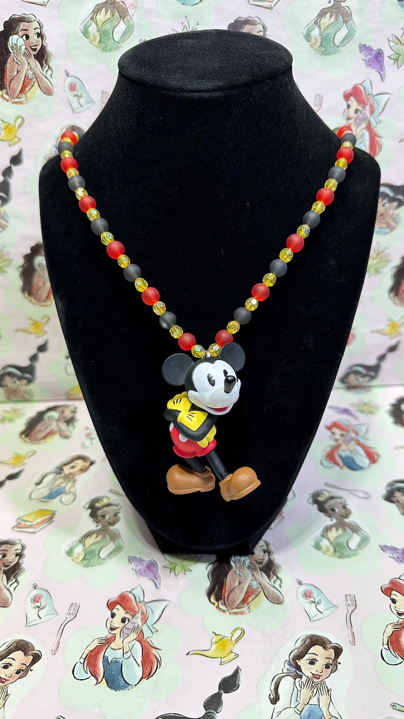 Puede incluir: Un collar con un colgante de Mickey Mouse. La cadena de cuentas tiene cuentas negras, rojas y amarillas. El colgante de Mickey Mouse est&aacute; en una pose de caminar, con pantalones cortos rojos, guantes amarillos y zapatos marrones.