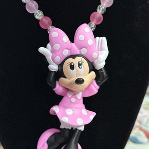 May include: A necklace featuring a Minnie Mouse pendant with a pink polka-dot dress and bow, white gloves, and black shoes. The chain is composed of alternating translucent clear beads and opaque pink beads, creating a colorful and playful accessory.