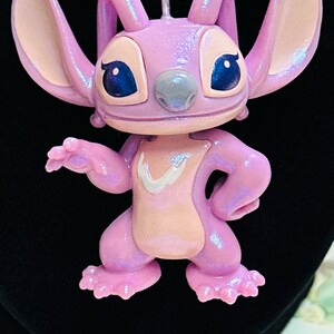 K&ouml;nnte beinhalten: Ein Anh&auml;nger in Form einer glitzernden, hellvioletten Figur mit gro&szlig;en Ohren, dunkelblauen Augen und einem hellrosa Brustfleck. Die Figur h&auml;ngt an einer Halskette aus runden, durchscheinenden violetten und rosafarbenen Perlen.