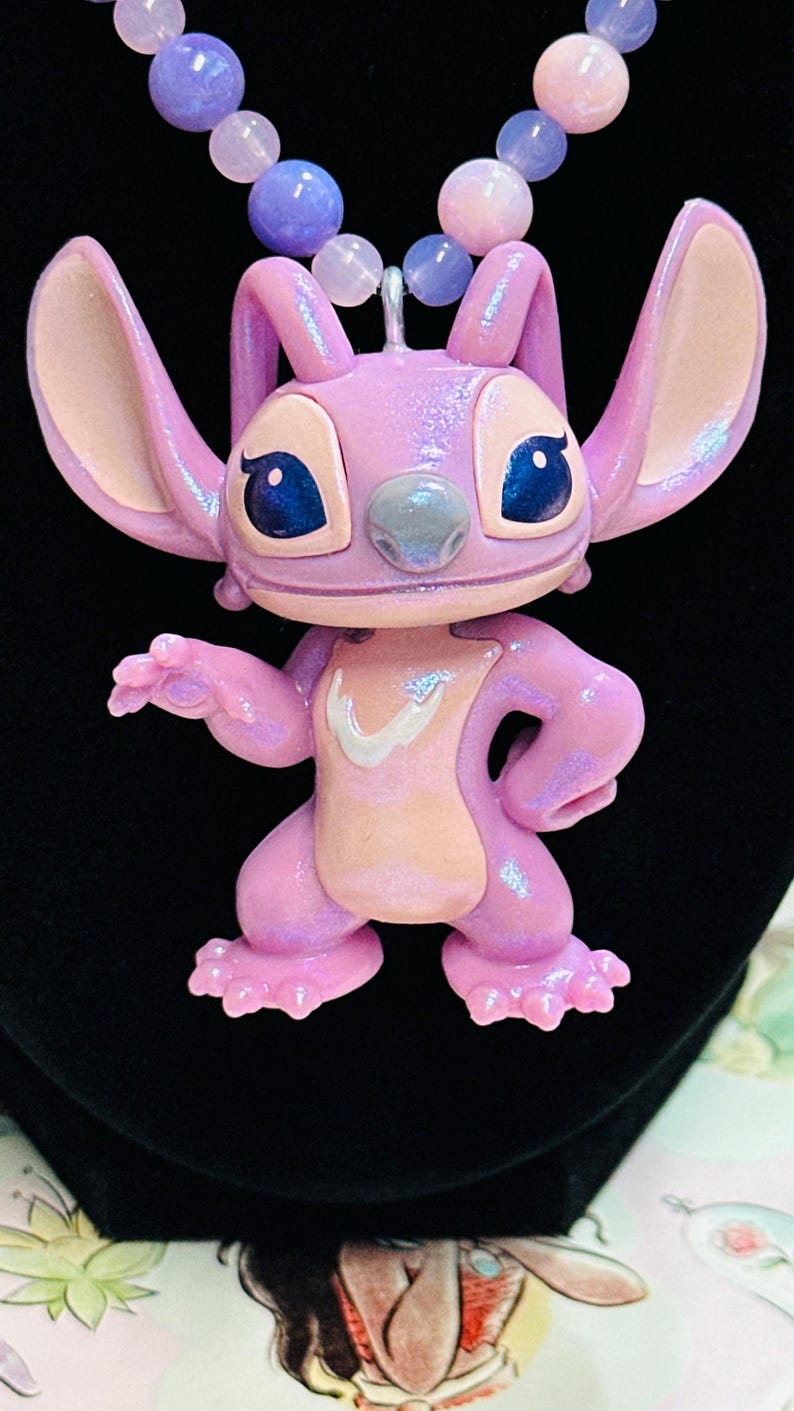 K&ouml;nnte beinhalten: Ein Anh&auml;nger in Form einer glitzernden, hellvioletten Figur mit gro&szlig;en Ohren, dunkelblauen Augen und einem hellrosa Brustfleck. Die Figur h&auml;ngt an einer Halskette aus runden, durchscheinenden violetten und rosafarbenen Perlen.