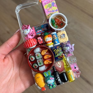 May include: A clear phone case decorated with numerous miniature food and character figurines, including a bowl of ramen, sushi, a box of Crunchy Pop cereal, a Takis bag, a Pringles can, M&Ms, a gumball machine, and several Hello Kitty figures.