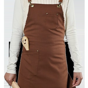 May include: A brown canvas apron with adjustable leather straps and a large front pocket. The apron has a wooden rolling pin and a wooden spoon with a heart-shaped face design in the pocket.