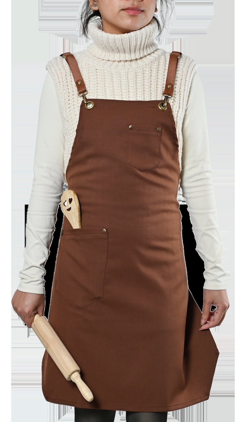 May include: A brown canvas apron with adjustable leather straps and a large front pocket. The apron has a wooden rolling pin and a wooden spoon with a heart-shaped face design in the pocket.