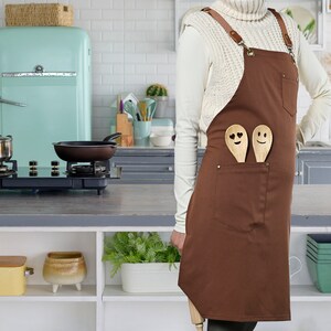 May include: A brown apron with two wooden spoons with smiley faces in the pocket. The apron has adjustable straps and a white sweater is worn over the apron.