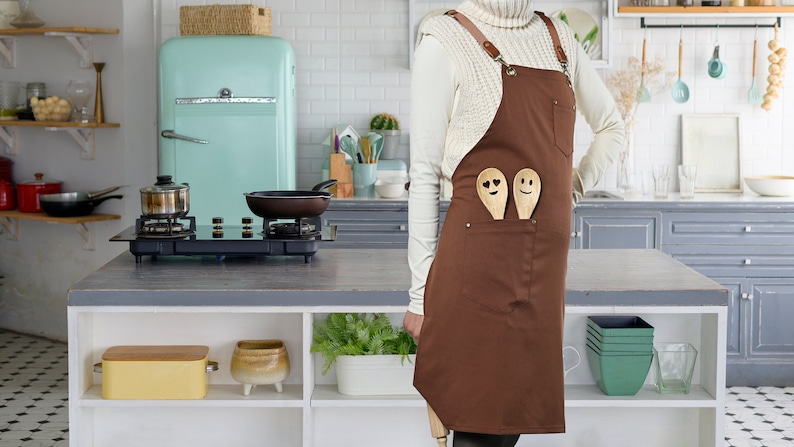 May include: A brown apron with two wooden spoons with smiley faces in the pocket. The apron has adjustable straps and a white sweater is worn over the apron.