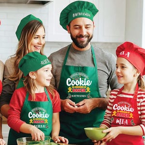 May include: A family of four wearing green and red Christmas-themed aprons with the text "Official Christmas Cookie Baker" and their names. The aprons have a gingerbread man design and holly leaves.