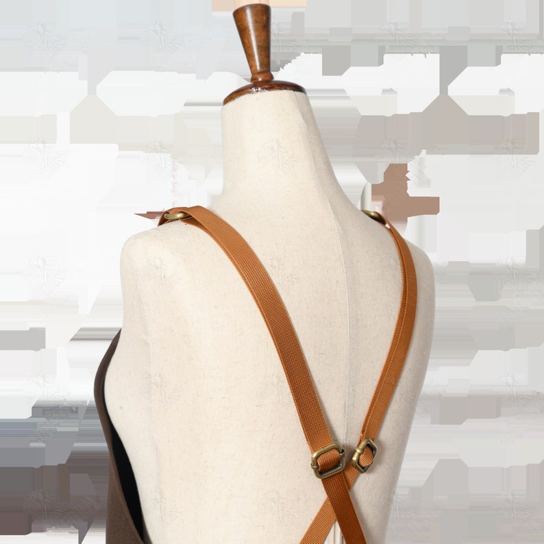 May include: Brown canvas apron with adjustable straps. The straps are made of a brown woven material and have metal buckles. The apron is shown on a white mannequin.