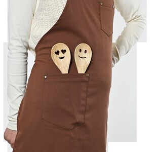 May include: Brown canvas apron with adjustable straps and a large front pocket. Two wooden spoons with smiley faces are in the pocket.
