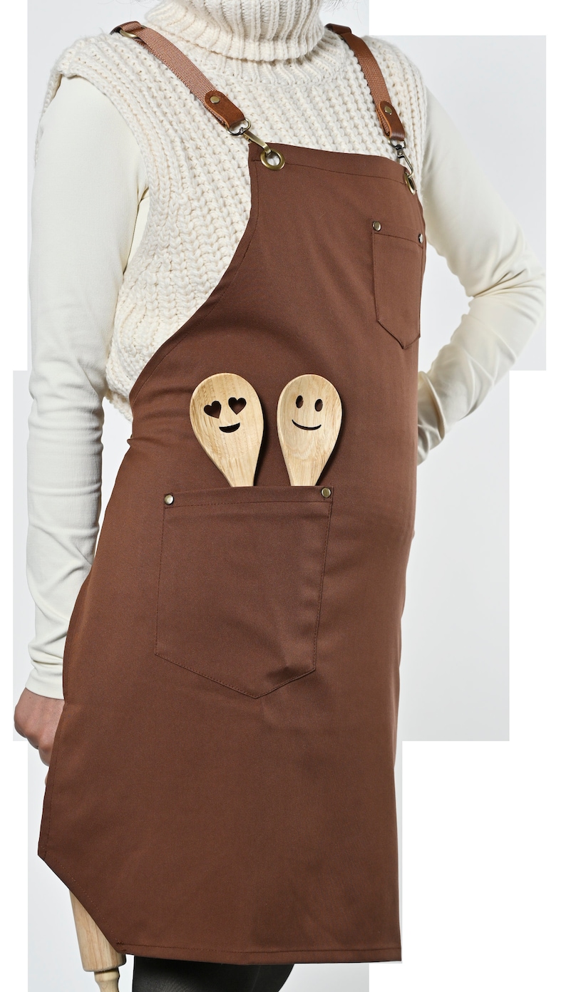 May include: Brown canvas apron with adjustable straps and a large front pocket. Two wooden spoons with smiley faces are in the pocket.