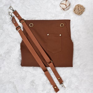 May include: A brown canvas apron with two adjustable straps and a single pocket. The apron has two brass eyelets on the top and is lying on a white fluffy surface.