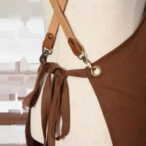 May include: Brown canvas apron with adjustable straps and a tie at the waist. The straps are made of a beige canvas material and have brown leather accents. The apron has a brass ring on the right side.