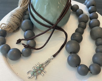 May include: A dark gray beaded necklace with a brown leather cord and a silver pendant with turquoise stones. The pendant is shaped like a stylized scorpion.