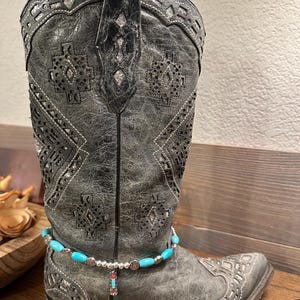Pode incluir: Um par de botas de cowboy cinzas com joias de tornozelo de contas turquesa e prata. As botas têm um padrão decorativo e estão usadas.