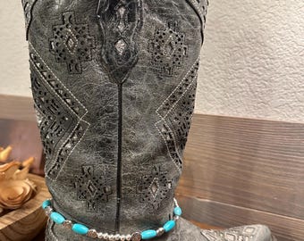May include: A pair of gray cowboy boots with turquoise and silver bead ankle jewelry. The boots have a decorative pattern and are worn.