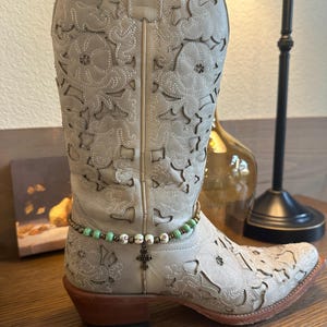 Pode incluir: Botas de cowboy bege com design floral e um bracelete de tornozelo com contas e um amuleto de cruz.