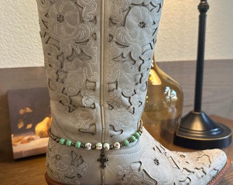 May include: Beige cowboy boots with a floral design and a beaded ankle bracelet with a cross charm.