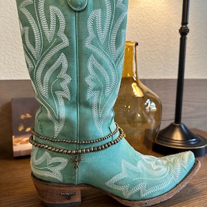 Pode incluir: Um par de botas de cowboy turquesa com costuras brancas e um amuleto de cruz marrom em um bracelete de tornozelo com contas.