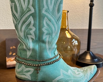 May include: A pair of turquoise cowboy boots with white stitching and a brown cross charm on a beaded ankle bracelet.