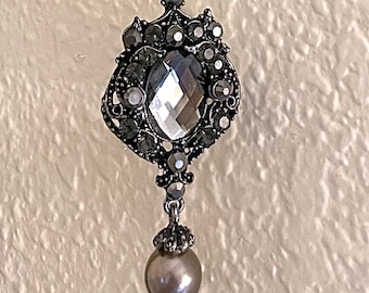 May include: A silver-toned metal pendant featuring a large, central faceted gray crystal surrounded by smaller dark gray crystals in an ornate setting, with a single round, smooth gray pearl-like bead dangling from the bottom.