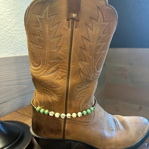 Pode incluir: Bota de cowboy de couro marrom com um bracelete de tornozelo de contas verdes e brancas.
