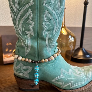 Pode incluir: Um par de botas de cowboy turquesa com costuras brancas e um bracelete de tornozelo com contas e pingentes de caveira turquesa.