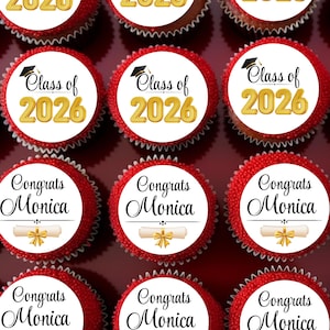 May include: A tray of red cupcakes with white frosting circles. Some cupcakes say "Class of 2026" with a graduation cap graphic. Others say "Congrats Monica" with a diploma graphic. The cupcakes are arranged in rows.