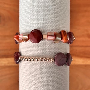 May include: Two bracelets with red and brown stone beads. The top bracelet has a mix of round and rectangular beads. The bottom bracelet has round beads and a rose gold chain.